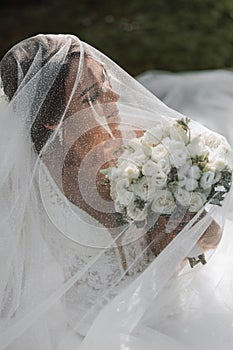 a bride in a wedding dress with a long train and a veil holds a wedding bouquet of roses while sitting under a veil