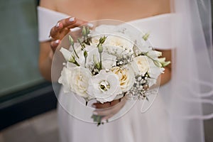 The bride in a wedding dress and a long veil holds a white wedding bouquet of roses close-up