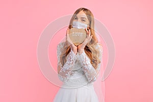 Bride in a wedding dress with a medical protective mask on her face, holding a wooden heart on a pink background. Quarantine,
