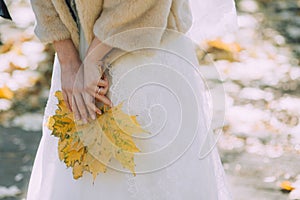 The bride in a wedding dress and a natural fur coat holds in hands yellow fallen leaves of a maple