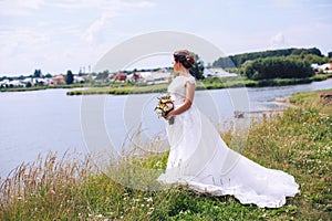 The bride in a wedding dress is standing on the river bank with a bouquet of flowers