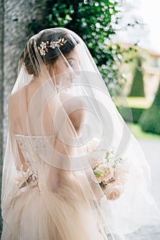 Bride in a wedding dress with a veil and a bouquet of flowers stands with her head turned near the wall. Lake Como. Back