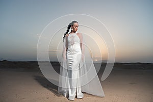 Bride in wedding gown on sunset sky. Woman in white dress in desert. Sensual woman with brunette hair. Fashion model in sand dunes