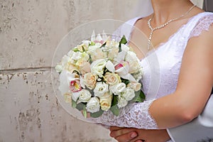 The bride in a white dress against a wall with a wedding bouquet, the groom embraces him from behind. hands of a man