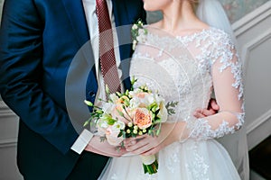 The bride in a white dress and groom in a blue suit are standing in the room and holding a wedding bouquet