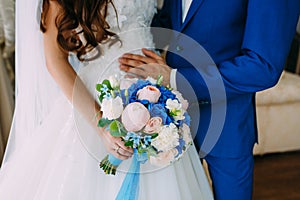 Bride in a white dress and groom in a blue tuxedo are standing next to the window and holding a wedding bouquet.