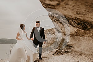 bride in white dress long veil and groom in suit walk in summer fall on mountain above river