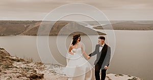 bride in white dress long veil and groom in suit walk in summer fall on mountain above river