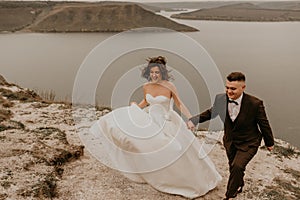 bride in white dress long veil and groom in suit walk in summer fall on mountain above river