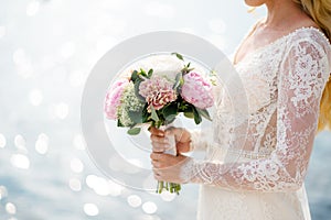 Bride in a white lace dress holds a bouquet of pink peonies in her hands