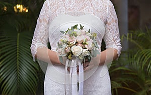 Bride in a white lace dress holds a bouquet of pink roses on a green foliage background