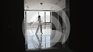 A bride in a white pantsuit and veil walks down a dark corridor to the window.