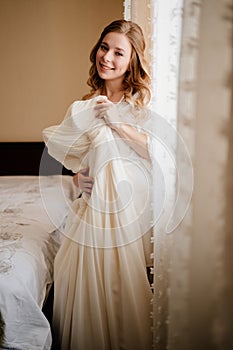 bride in a white silk robe with the wedding dress by the window