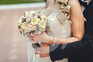 The bridegroom embraces the bride`s waist, a wedding bouquet in her hands, close-up, in focus a bouquet, a blurry background