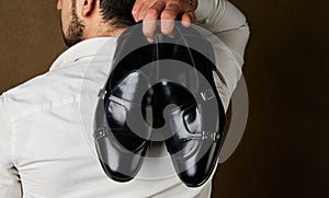 Bridegroom holding elegant black shoes behind his back over brown background