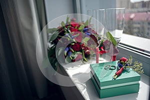 Brides bouquet of red roses before the wedding with rings and wine glasses stands on the window overlooking the street
