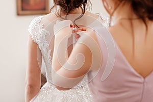 Bridesmaid helping bride fasten corset and getting her dress, preparing bride in morning for the wedding day