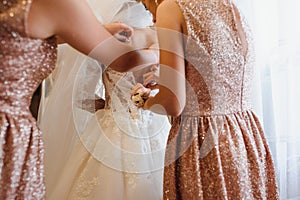 Bridesmaid helping slender bride lacing her wedding white dress, buttoning on delicate lace pattern with fluffy skirt on waist.
