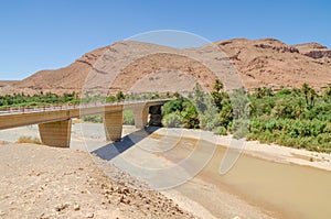 Bridge spanning over dry river bed with some water, mountains and palms in Morocco, North Africa