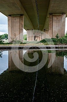 A bridge from underneath reflected in water. A dividing line between the two carriageways creates a stark white line