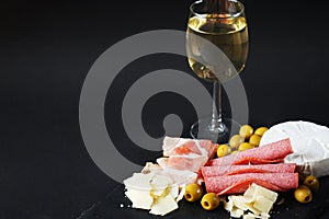 Brie cheese with salami, bacon, parmesan, olives next to a glass of white wine on a tray