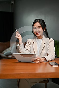 Bright Asian businesswoman at her desk, using tablet, smiling and looking at the camera