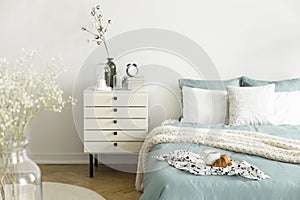 A bright bedroom interior with sage green and white bedding, pillows on bed and a drawer nightstand. Real photo.