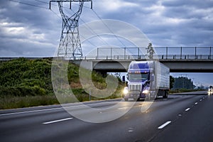 Bright blue big rig semi truck transporting cargo in refrigerated semi trailer running under the bridge on the wide highway road