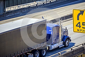 Bright blue big rig semi truck transporting commercial cargo in covered semi trailer with frame driving on the interstate highway