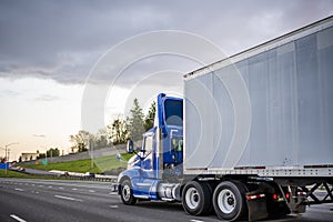 Bright blue day cab big rig semi truck with roof spoiler transporting cargo in dry van semi trailer running on the evening road