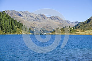 Bright blue mountain lake under blue sky with woods, stones and snow