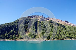 Bright blue mountain lake under blue sky with woods, stones and snow