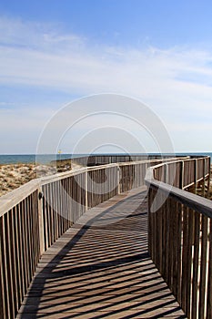 Bright blue skies over long wooden pier that stretches out over sandy dunes, leading one to the beach beyond