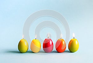 Bright burning paraffin candles in the shape of colorful Easter eggs on blue background