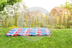 Bright checkered mat on green grass lawn under trees in garden. Blurred wooden houses on background. Empty space for