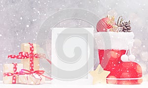 Bright christmas mock up with photo frame: festive gift boxes, toys, and fir-cones in red santa`s boot under snow