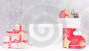 Bright christmas mock up with photo frame: festive gift boxes, toys, and fir-cones in red santa`s boot under snow