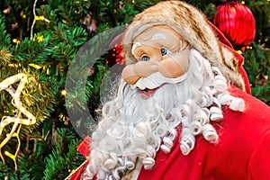Bright colorful Santa Claus doll in red costume with long white beard on green fir tree branches background. Christmas