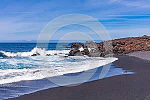The bright contrast of the white sea foam of the waves rolling onto the shore with the sand and black solidified lava