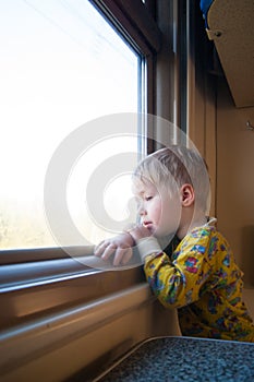 A bright, cute three-year-old boy riding a train looks out the window, behind which a dull landscape sweeps through