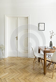 A bright dining room interior with a wooden table and chairs standing on a herringbone parquet against a white wall with double do