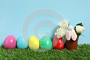 Bright Easter eggs, pot with flowers and toy bunny on green grass against light blue background