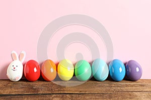Bright Easter eggs and white one as bunny on wooden table against pink background, space for text