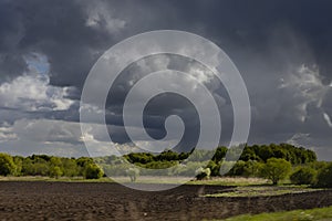 Bright epic thunderstorm landscape in early spring with dark blue fluffy clouds in sunlights above black field arable land.