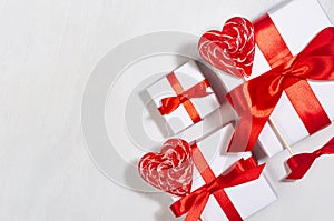 Bright festive wedding background  - white gift boxes with red bow, sweet lollipops hearts on white wood board, border, closeup.