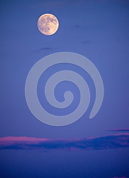 Bright full moon in early evening sky with long, pink-tinged cloud bank below