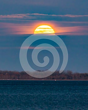 Bright full moon glowing in a hazy night sky popping out from behind clouds on the horizon.