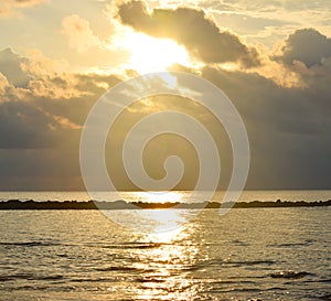Bright Golden Yellow Sun behind Cloud and Sunbeams, Reflection of Sunlight on Sea Water, Dark Clouds, and Warm Colors in Sky