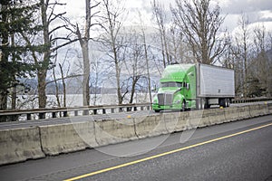 Bright green big rig semi truck transporting frozen cargo in refrigerator semi trailer running on the highway road along the river
