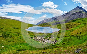 Bright green blue alpine landscape with mountain lake in highland valley in sunlight and big mountain under blue cloudy sky.